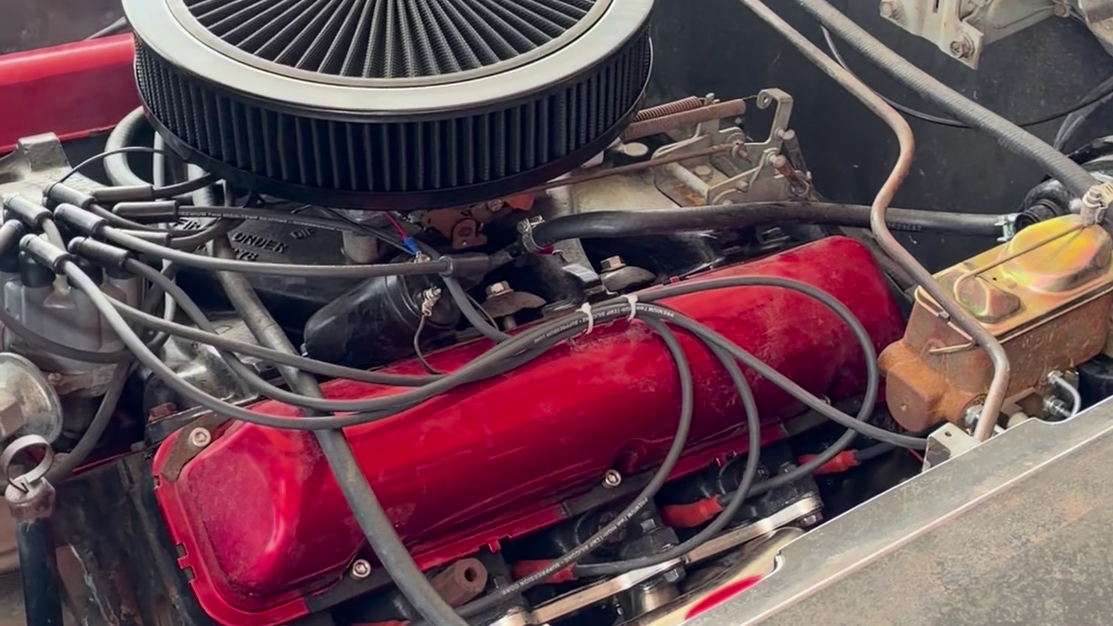 A carbureted V8 running with the hood up in the shop